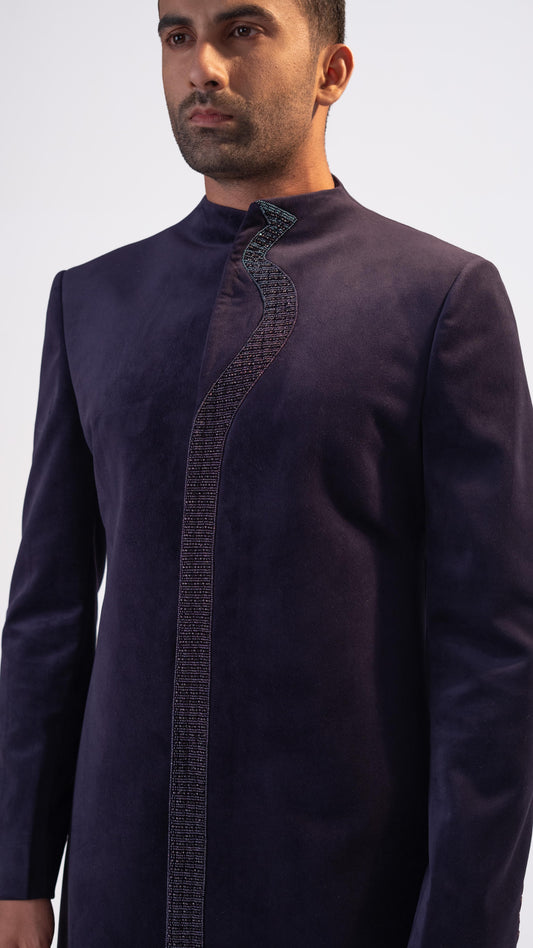 A man wearing a dark purple long-sleeve indowestern with a textured vertical embroidered placket and a high stand collar, photographed against a light gray studio background.
