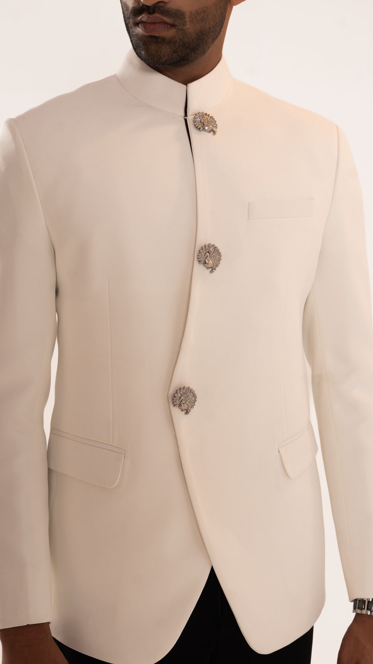 A close-up of a man wearing a cream-colored traditional Indian jacket (bandhgala) with ornate silver-toned decorative brooches, paired with black trousers against a plain white background.