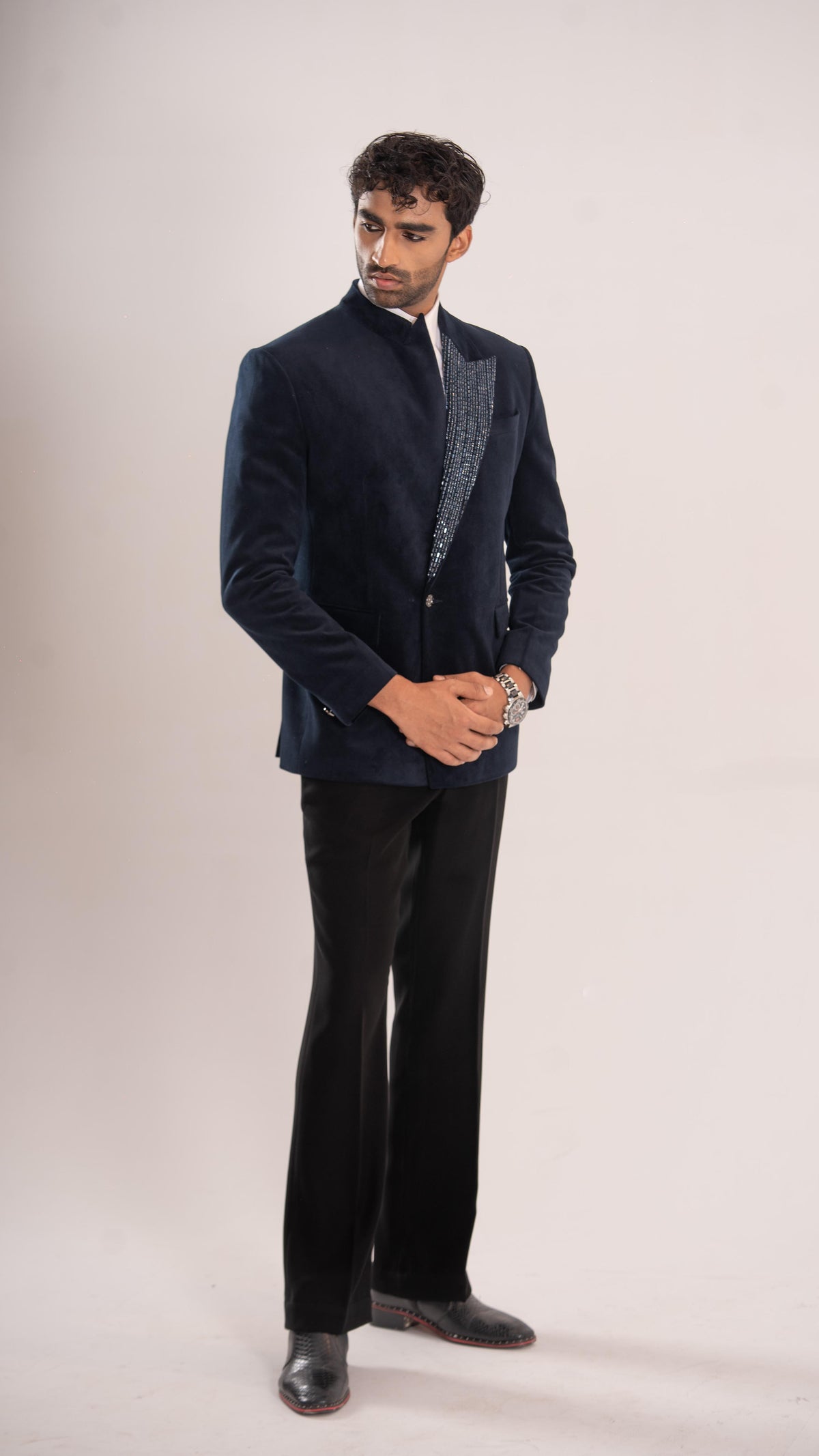A man modeling a dark navy blue velvet blazer with textured silver-gray lapels, paired with black trousers and black leather shoes, standing against a plain light beige studio background.