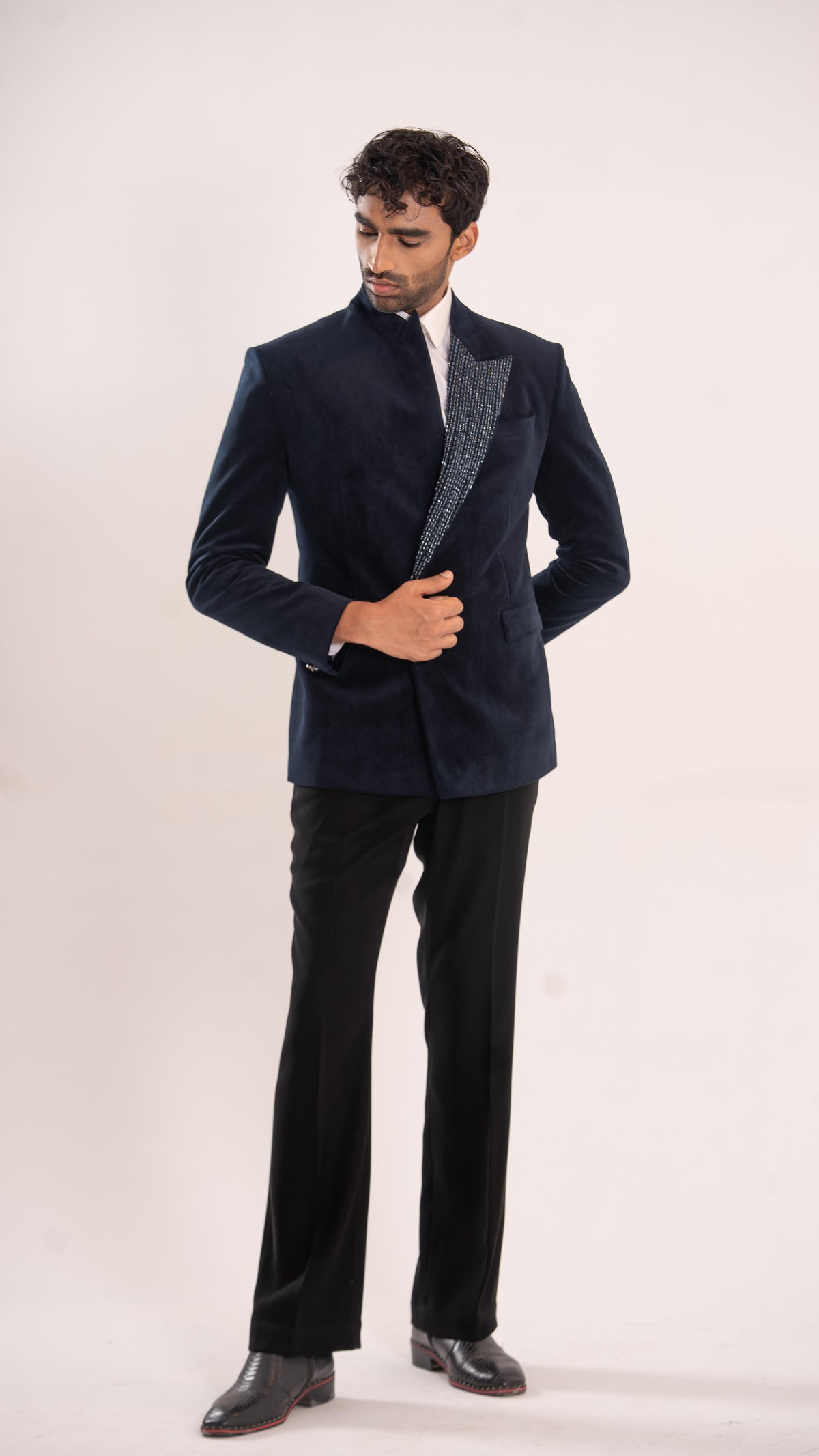 A man modeling a dark navy blue velvet blazer with textured gray lapels, paired with black trousers and black dress shoes, against a plain light background.