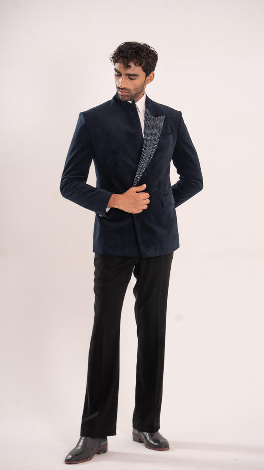 A man modeling a dark navy blue velvet blazer with textured gray lapels, paired with black trousers and black dress shoes, against a plain light background.