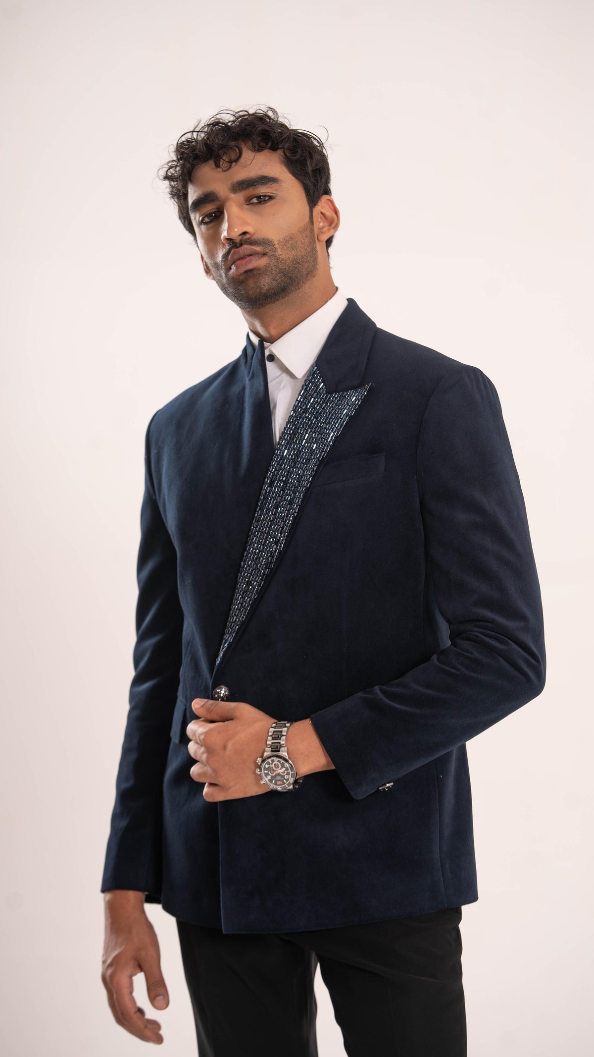 A man wearing a navy blue velvet blazer with textured, patterned lapels, a white dress shirt, and a dark tie, standing against a plain light background.