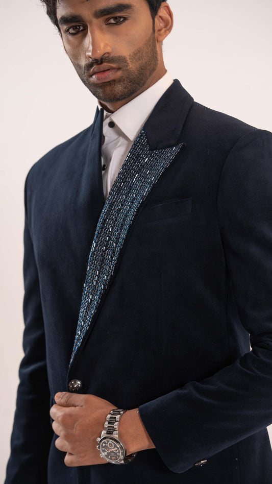 A man wearing a dark navy velvet blazer with a sparkling silver and blue beaded lapel detail, paired with a white dress shirt and a silver watch.