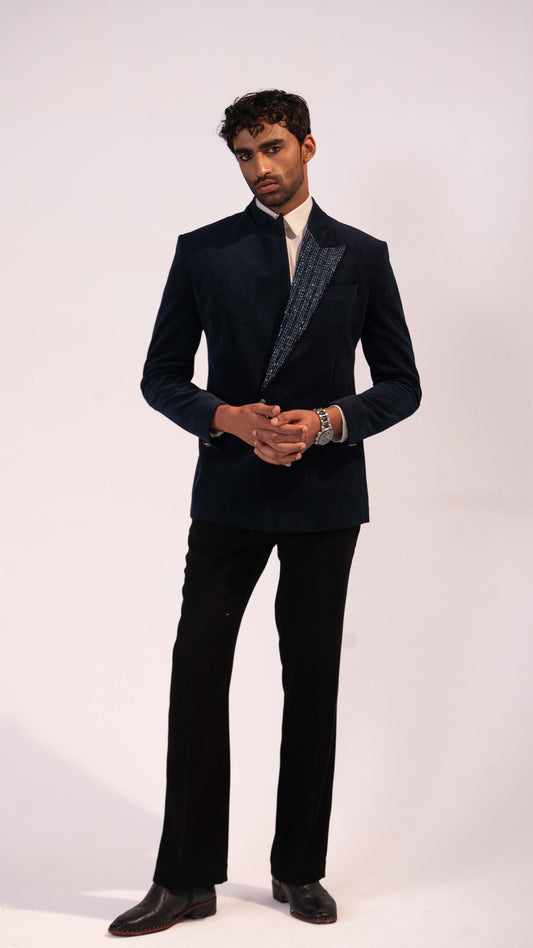 A man modeling a dark navy blue velvet blazer with textured patterned lapels, paired with black trousers and black dress shoes, against a plain light background.