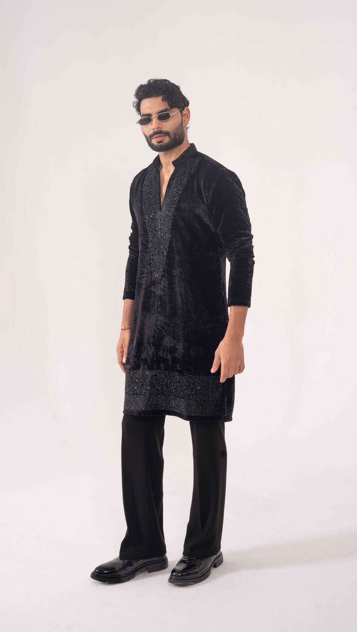 A man modeling a black velvet long kurta with subtle silver embroidery, paired with black trousers and black leather loafers, set against a plain white studio background.