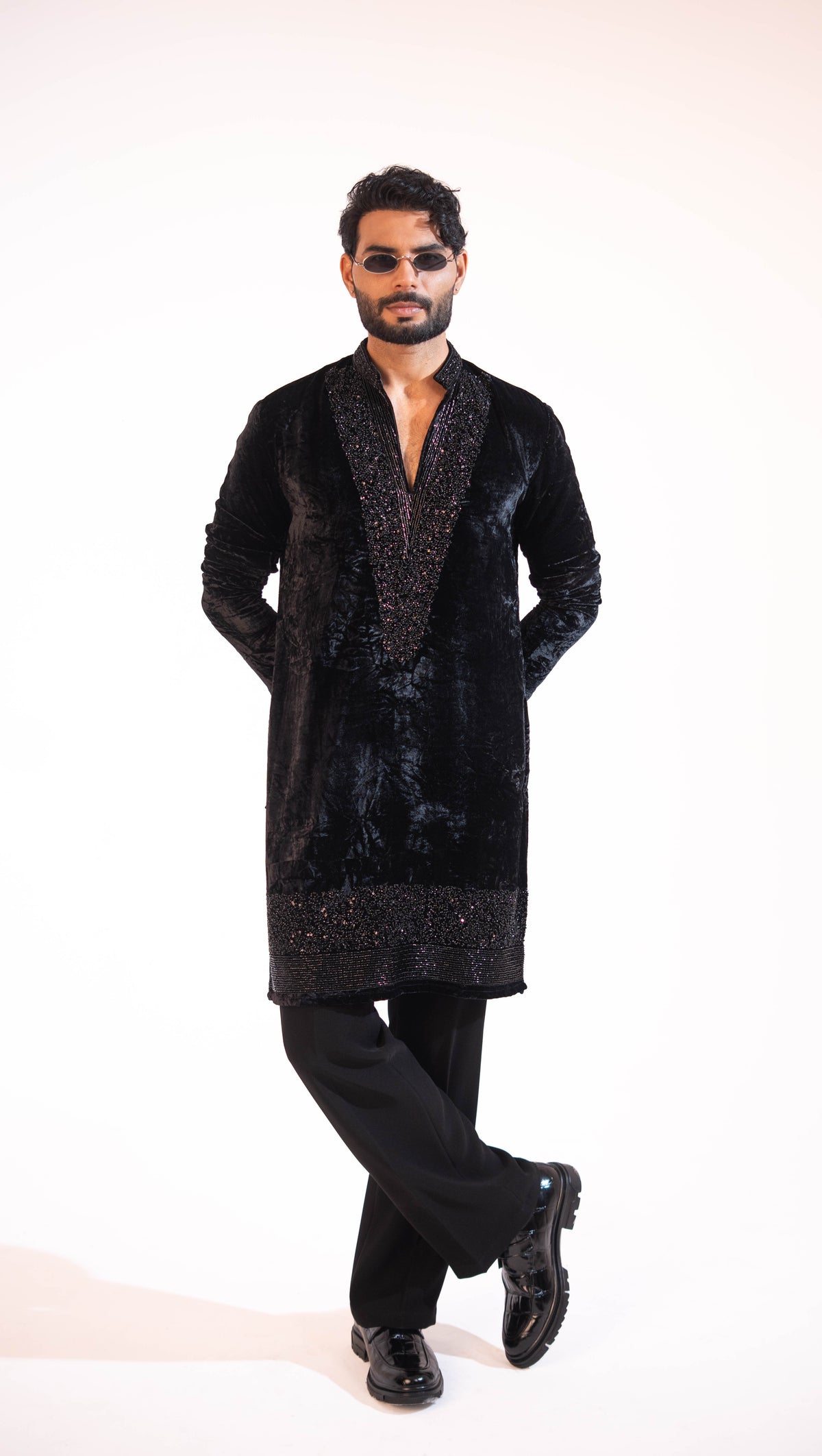 A man modeling a black velvet long kurta with textured metallic embroidery on the front panel and hem, paired with black trousers and black shoes, against a plain white studio background.
