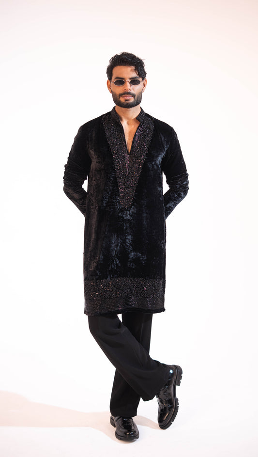 A man modeling a black velvet long kurta with textured metallic embroidery on the front panel and hem, paired with black trousers and black shoes, against a plain white studio background.