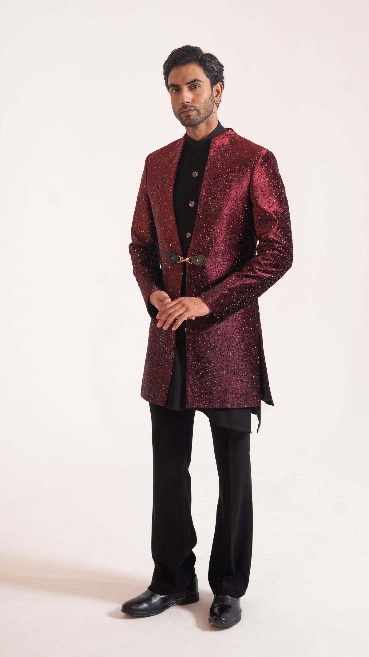 A man modeling a metallic burgundy sherwani jacket with a gold-tone belt over a black kurta and black trousers, standing against a plain off-white studio background.