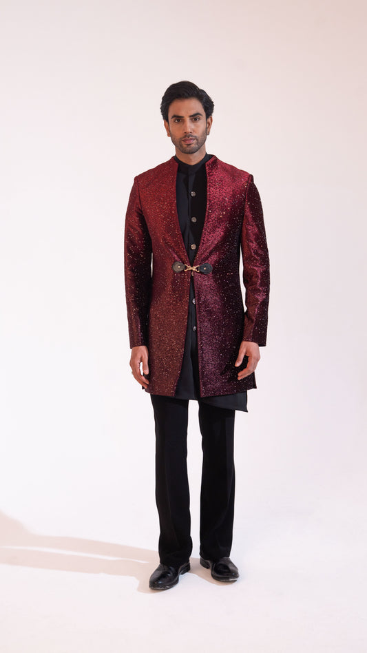 A man modeling a textured, metallic deep red sherwani jacket over a black kurta and black trousers, standing against a plain off-white studio background.