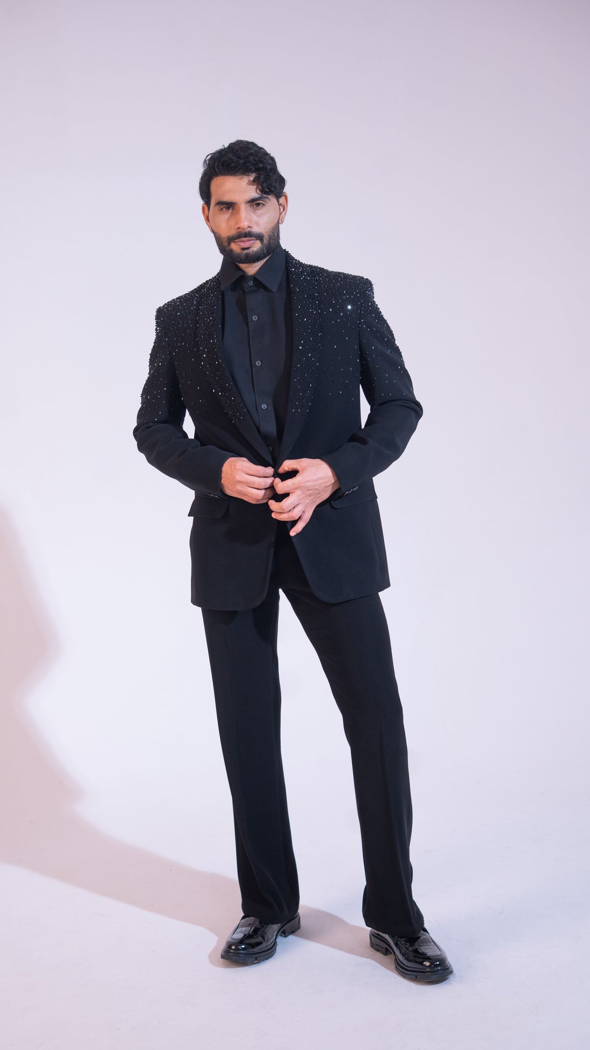 A man modeling a black formal suit with sparkling embellishments on the jacket, paired with a black shirt and trousers, standing against a plain light background.