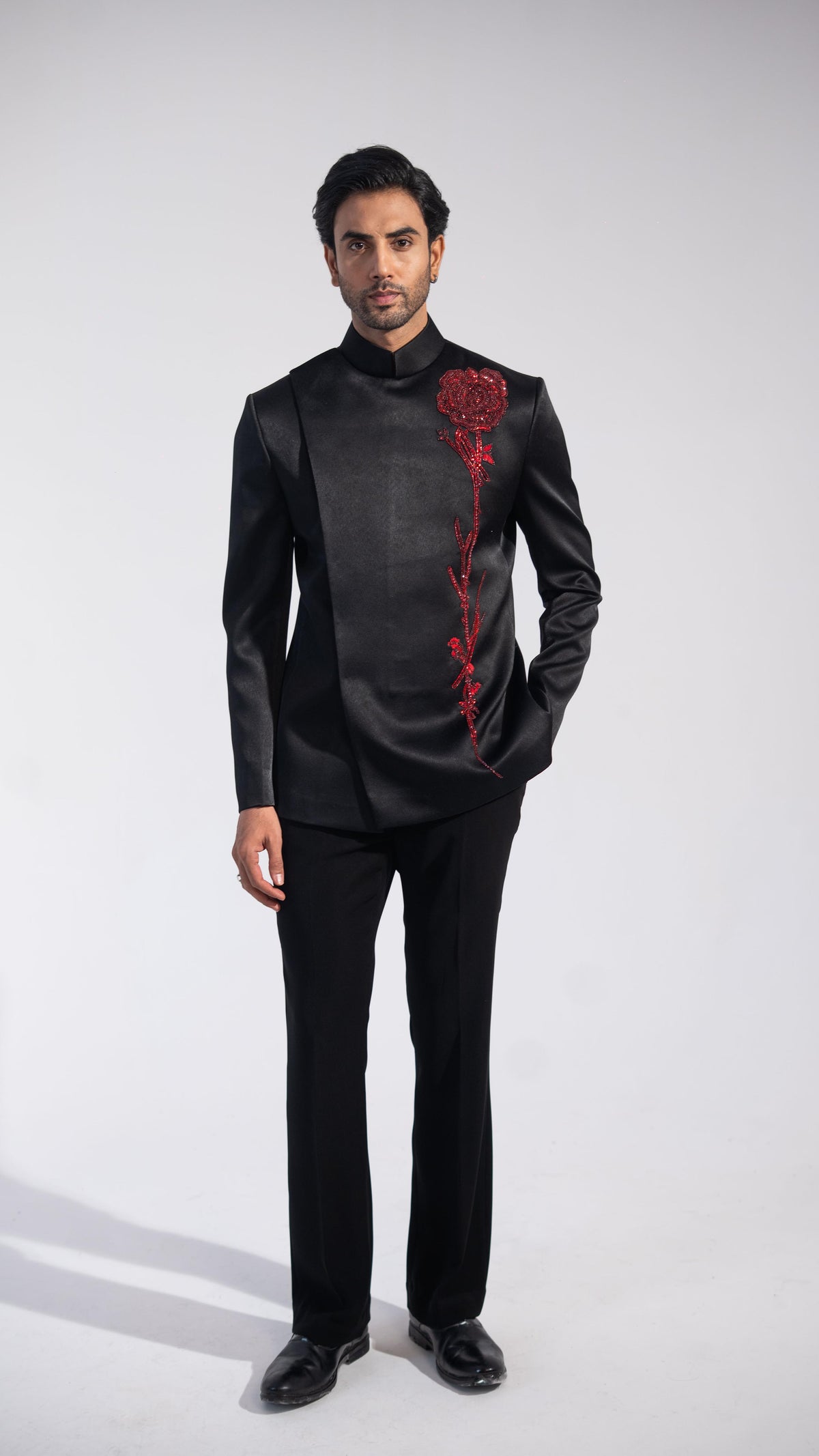 A man modeling a black satin-like traditional suit jacket with a high collar and a red rose embroidery on the chest, paired with black trousers and black shoes, against a light gray studio background.