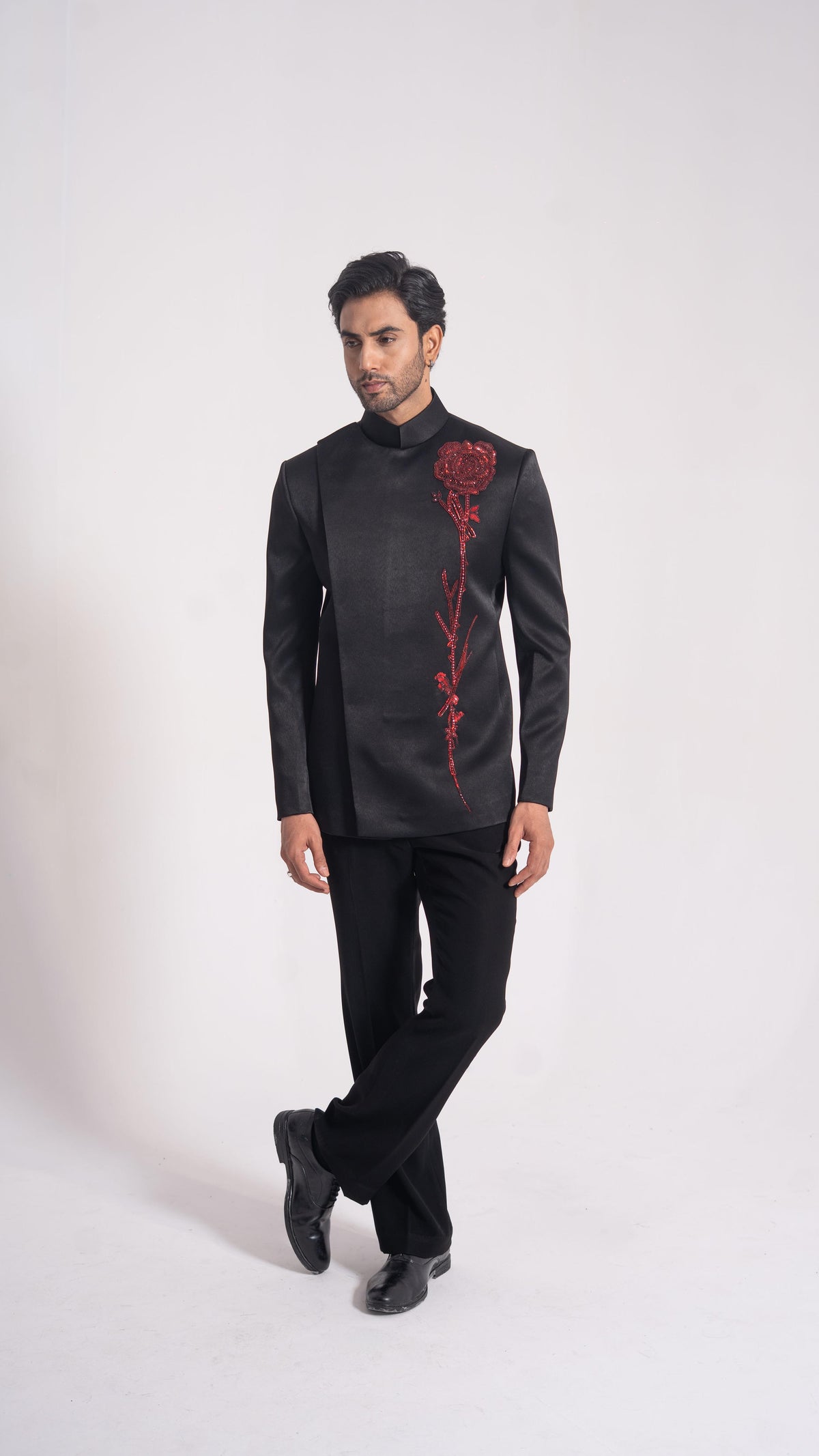 A man modeling a black traditional Indian jacket with a red rose embroidery on the front, paired with black trousers and black shoes, against a plain light gray studio background.