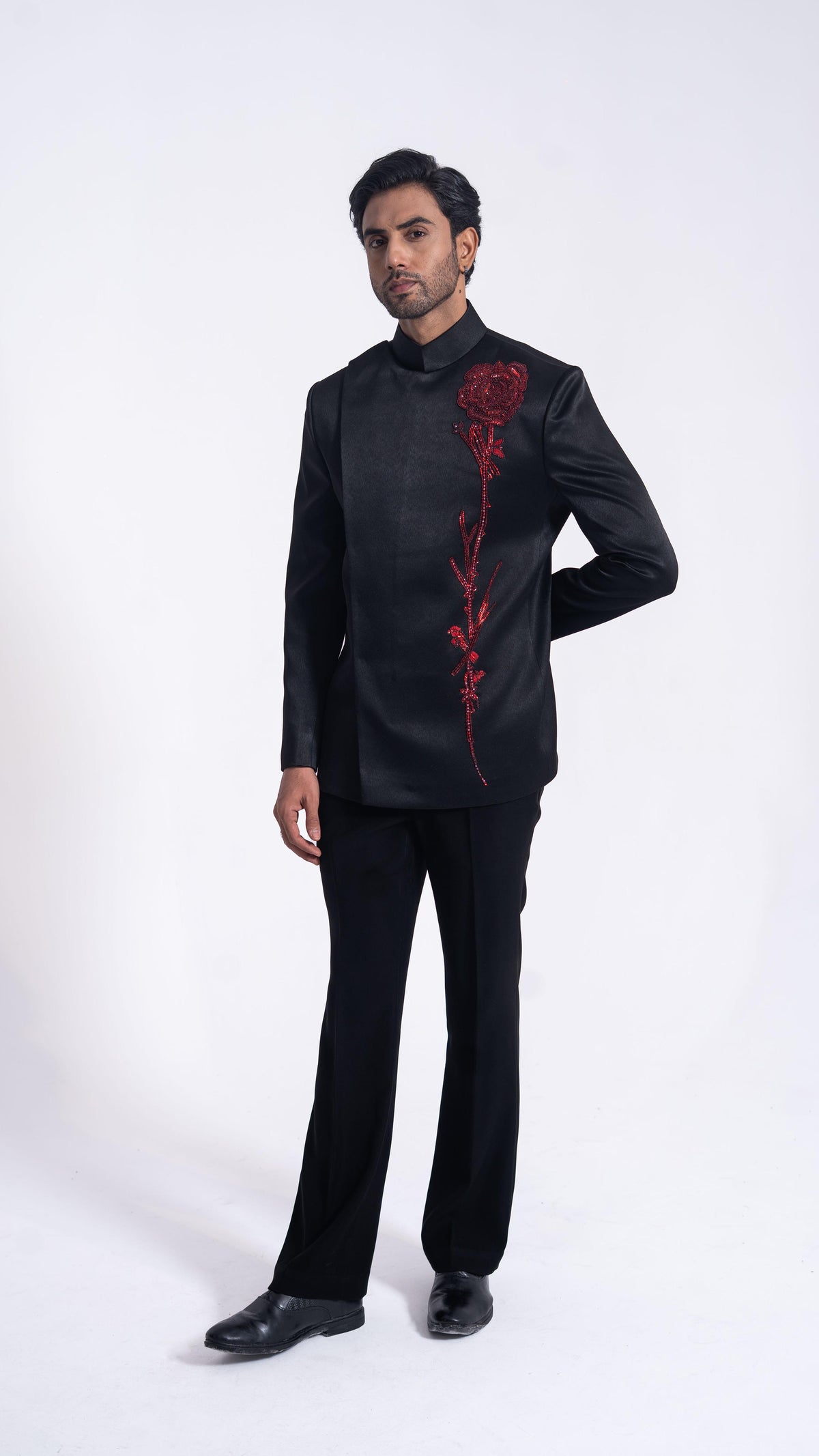 A man modeling a black long-sleeve traditional jacket with a red rose embroidery on the chest, paired with black trousers, against a plain white studio background.