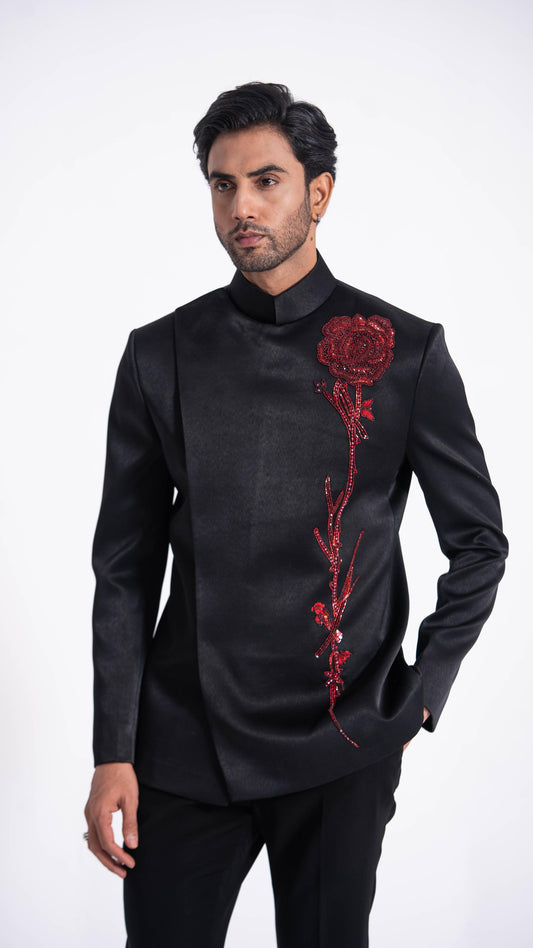 A man wearing a black satin Nehru jacket with a red rose embroidery on the chest, paired with black trousers, against a plain white studio background.