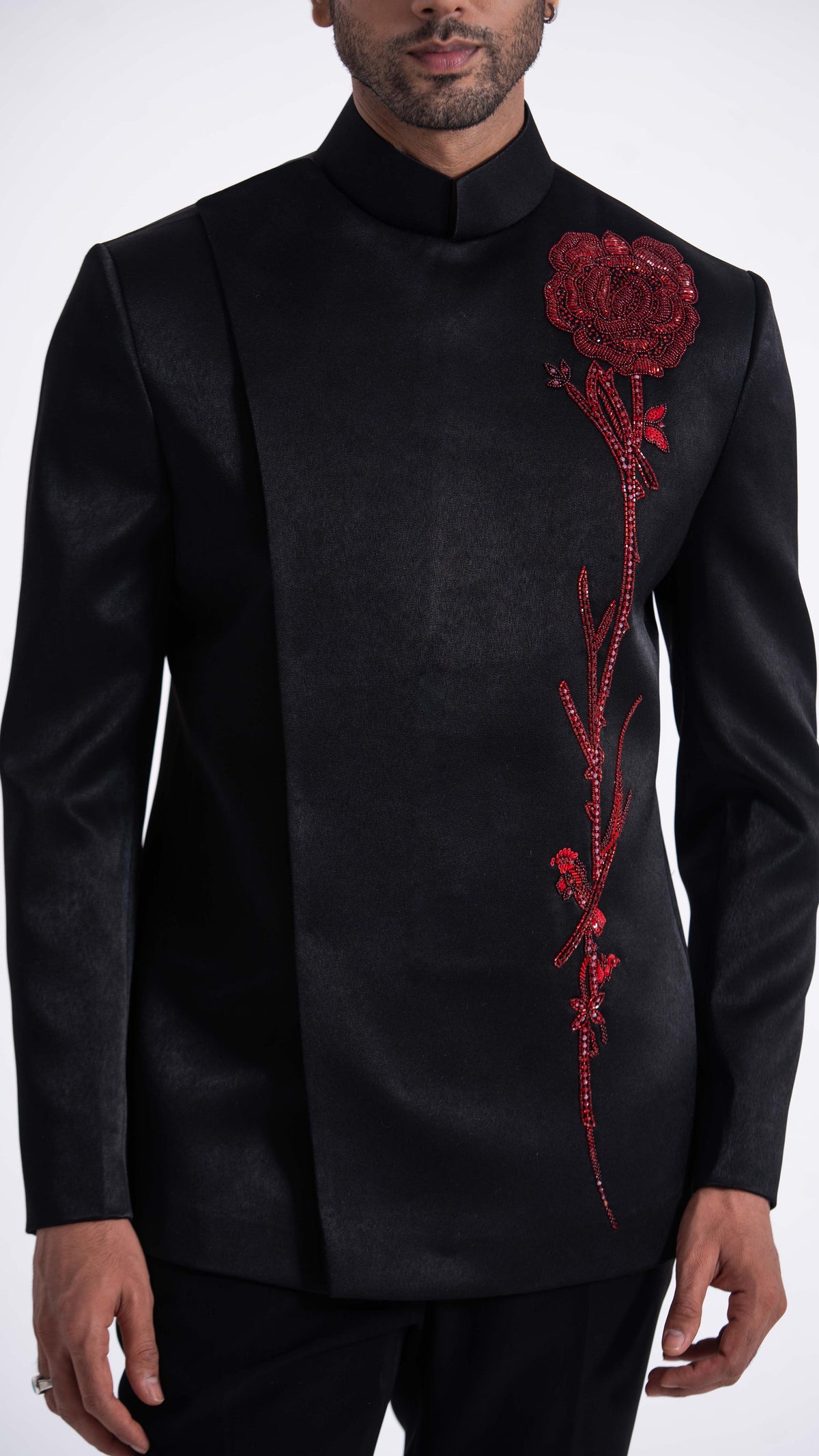 A man wearing a black formal jacket with a red embroidered rose motif on the chest, photographed against a plain light background.