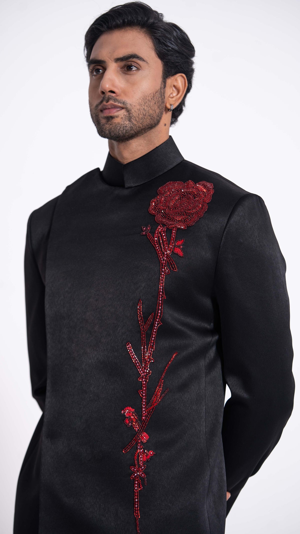 A man wearing a black high-neck traditional jacket with a large red rose embellishment on the chest, photographed against a plain light gray background.