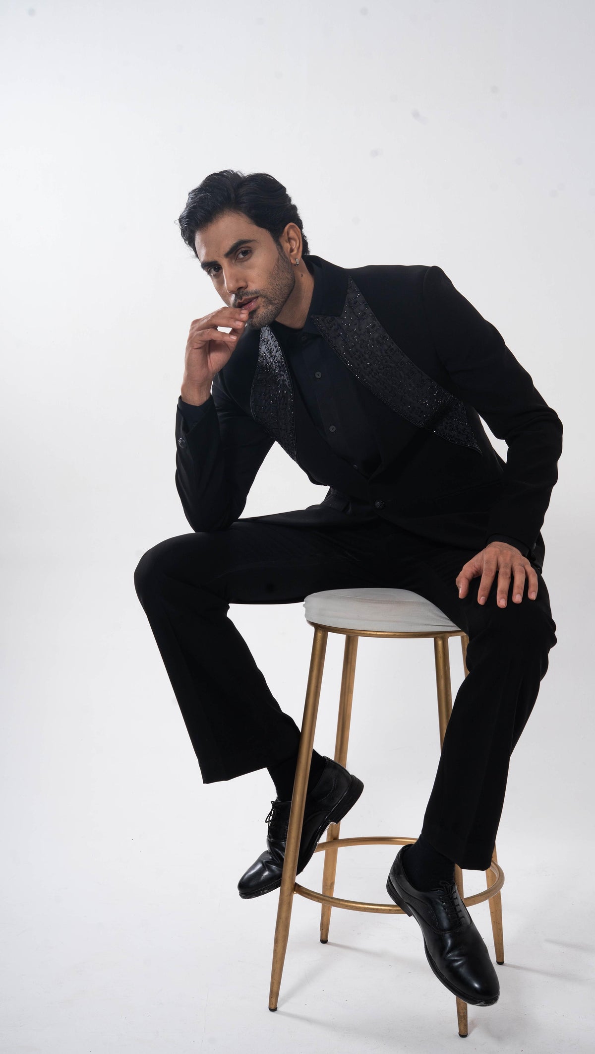 A man in a black suit with a patterned lapel vest and black dress shoes sits on a gold-framed stool with a white cushion against a plain white studio background.
