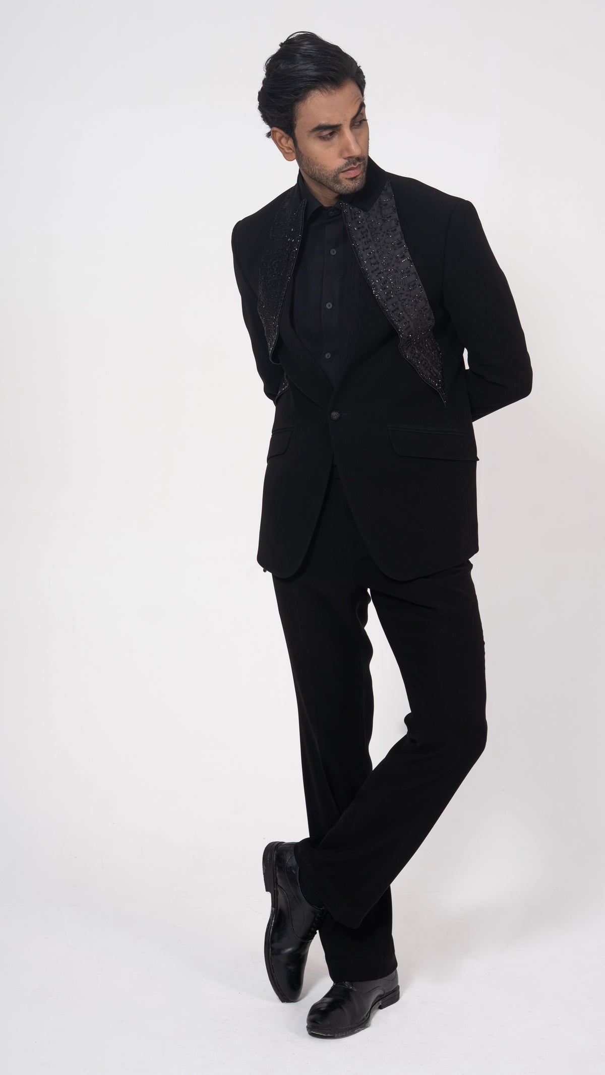 A man modeling a black formal suit with a black shirt, a textured black necktie, and black dress shoes, standing against a plain white studio background.
