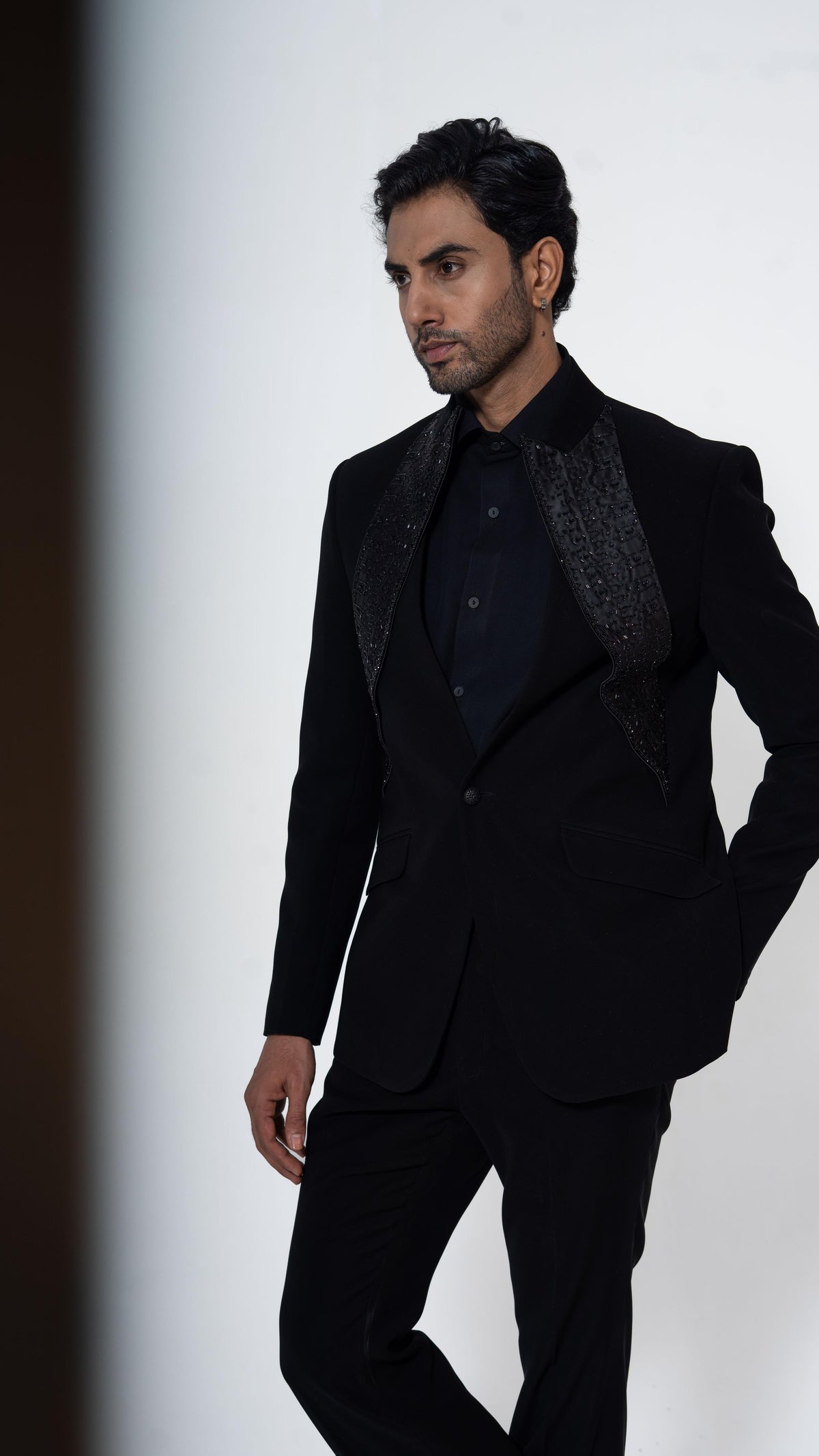 A man modeling a black formal suit with textured patterned lapels, a black shirt, and black trousers against a clean white studio background.