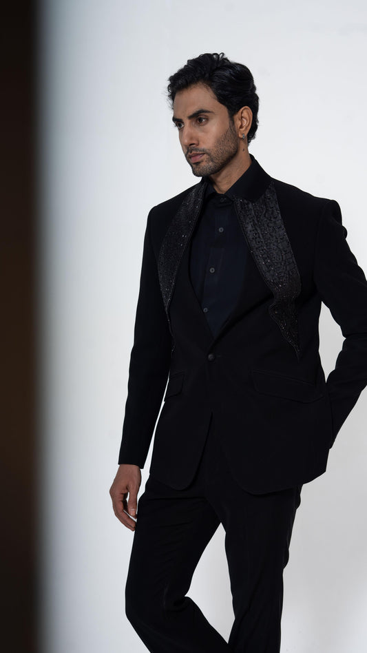 A man modeling a black formal suit with textured patterned lapels, a black shirt, and black trousers against a clean white studio background.
