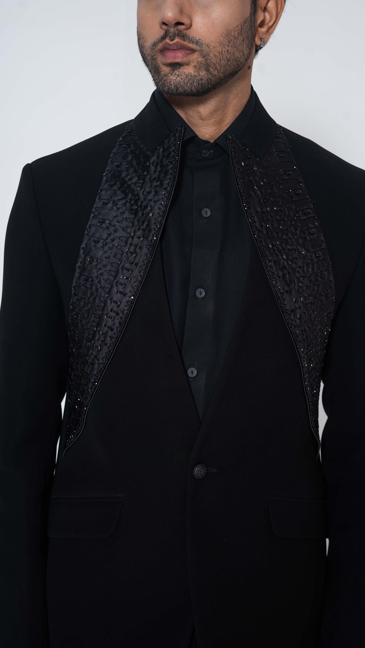 A close-up of a man wearing a black formal jacket with textured, patterned shoulder panels over a black shirt, against a plain light gray background.