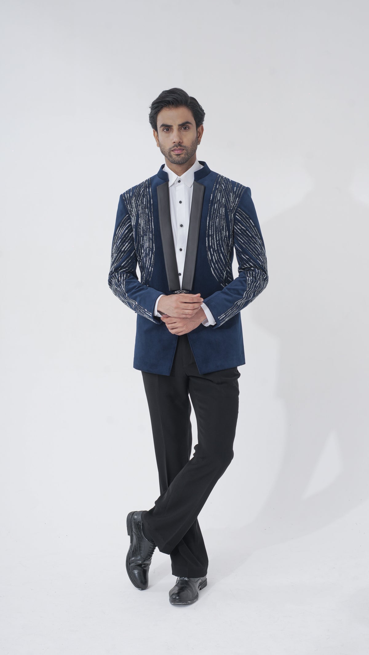 A man modeling a dark blue tuxedo with textured silver patterns on the sleeves and lapels, paired with black trousers, a white shirt, and black dress shoes, against a plain light gray background.