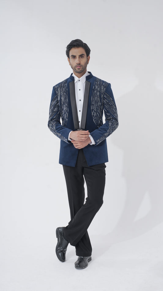 A man modeling a dark blue tuxedo with textured silver patterns on the sleeves and lapels, paired with black trousers, a white shirt, and black dress shoes, against a plain light gray background.