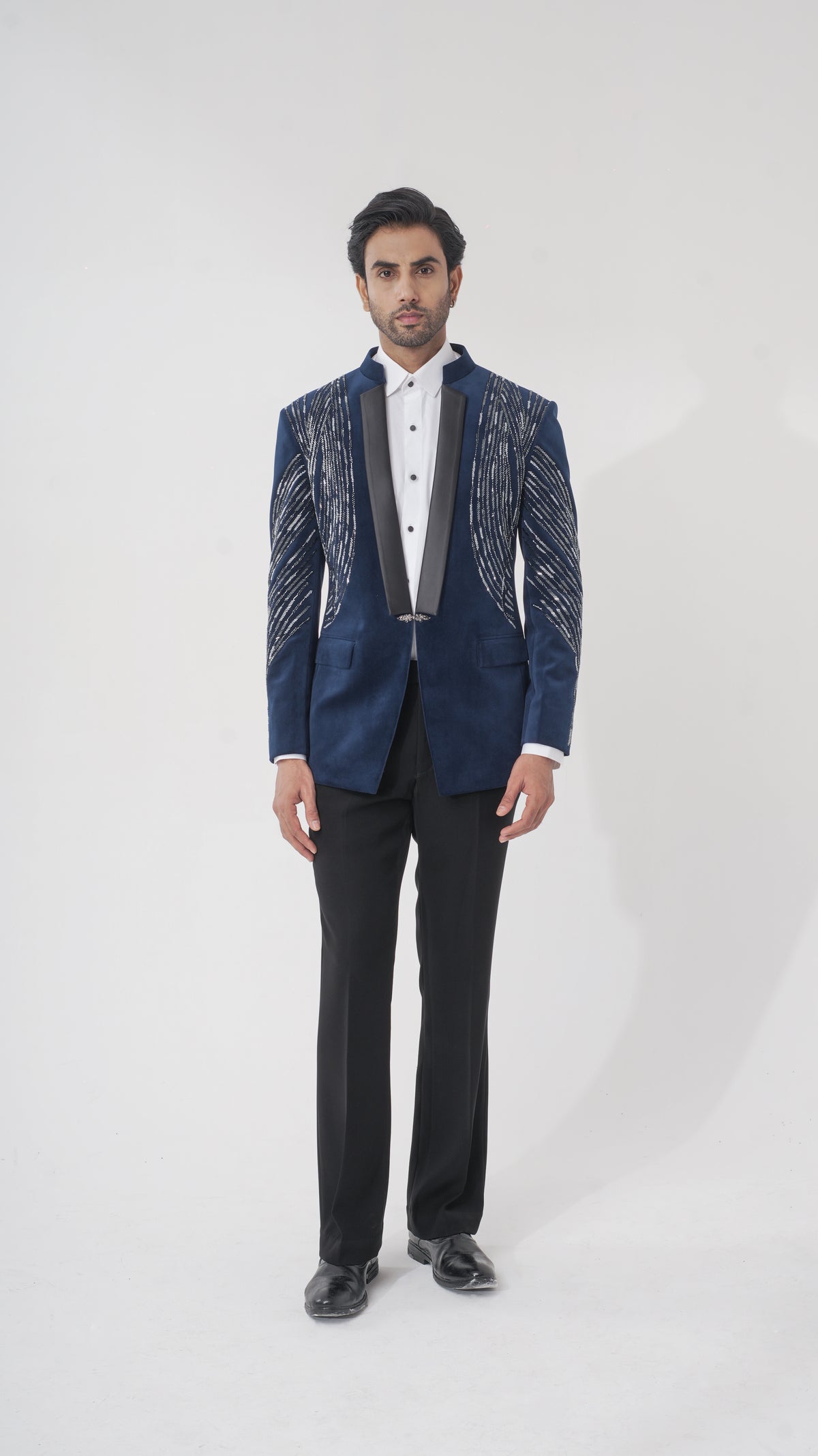 A man modeling a dark blue formal tuxedo with silver metallic embroidered patterns on the shoulders and sleeves, paired with a white shirt, black trousers, and black dress shoes, against a light gray studio background.
