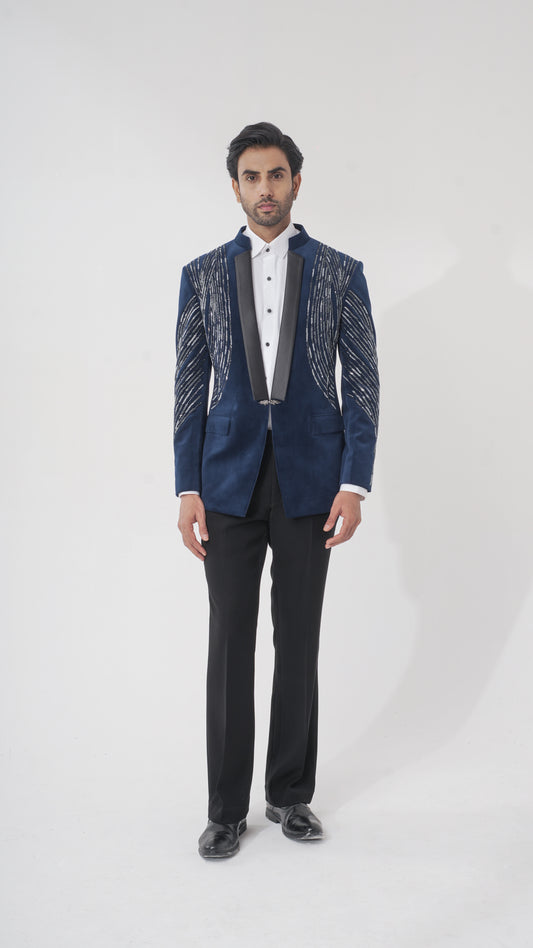 A man modeling a dark blue formal tuxedo with silver metallic embroidered patterns on the shoulders and sleeves, paired with a white shirt, black trousers, and black dress shoes, against a light gray studio background.