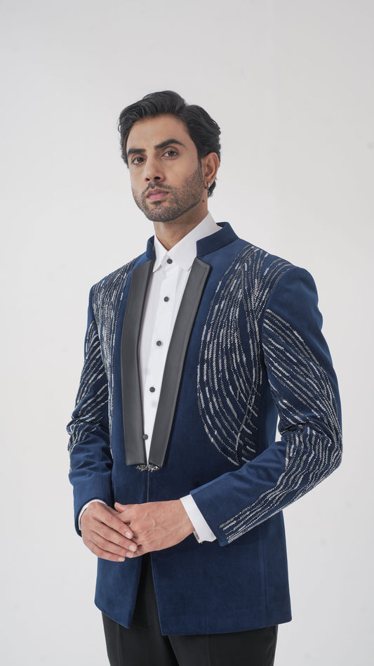 A man wearing a navy blue velvet tuxedo with silver metallic embroidered patterns on the sleeves and a black satin peak collar, paired with a white shirt and black trousers, against a light gray studio background.