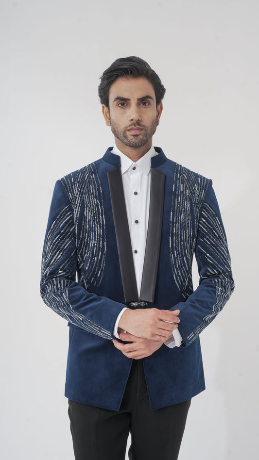 A man wearing a navy blue velvet tuxedo with silver embroidered patterns on the shoulders and sleeves, paired with a white shirt and black trousers, standing against a light gray background.