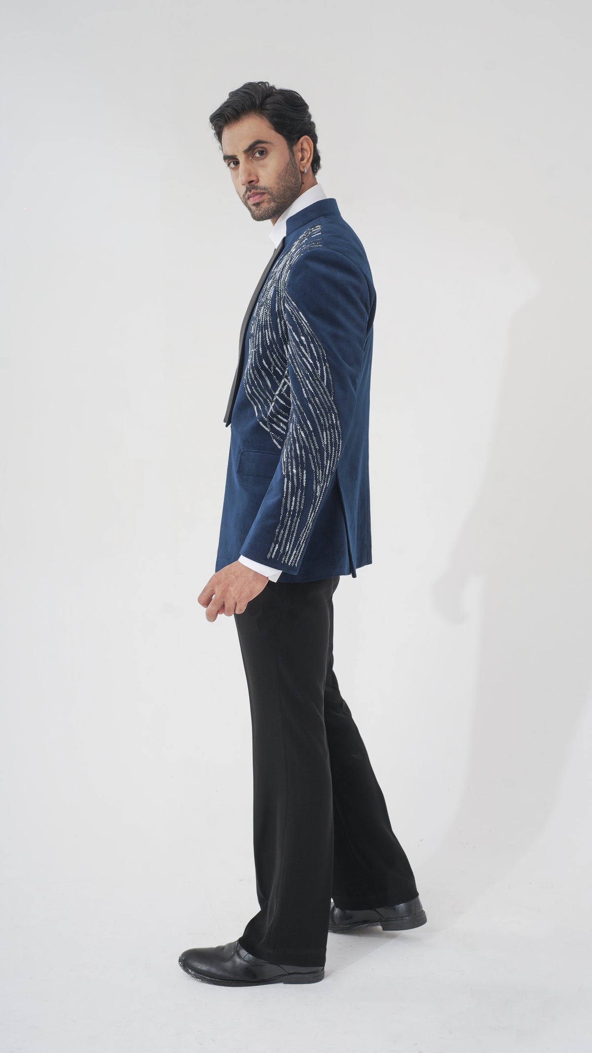 A man modeling a navy blue tuxedo with intricate silver and white embroidered patterns on the sleeves, paired with black trousers and black dress shoes, against a plain white studio background.