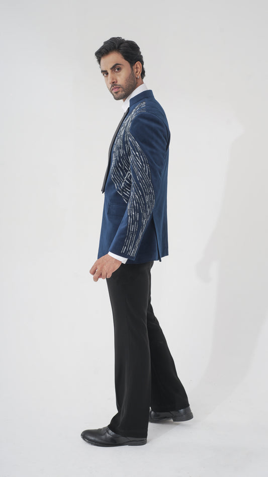 A man modeling a navy blue tuxedo with intricate silver and white embroidered patterns on the sleeves, paired with black trousers and black dress shoes, against a plain white studio background.