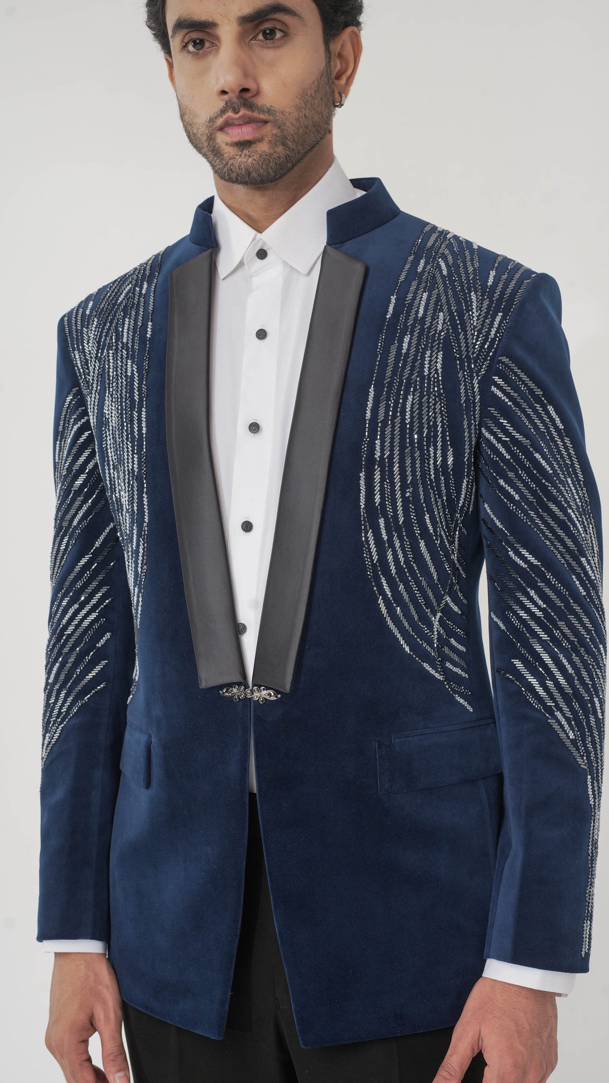 A man wearing a navy blue velvet tuxedo with silver embroidered patterns on the shoulders and sleeves, paired with a white dress shirt and black trousers, against a light gray background.