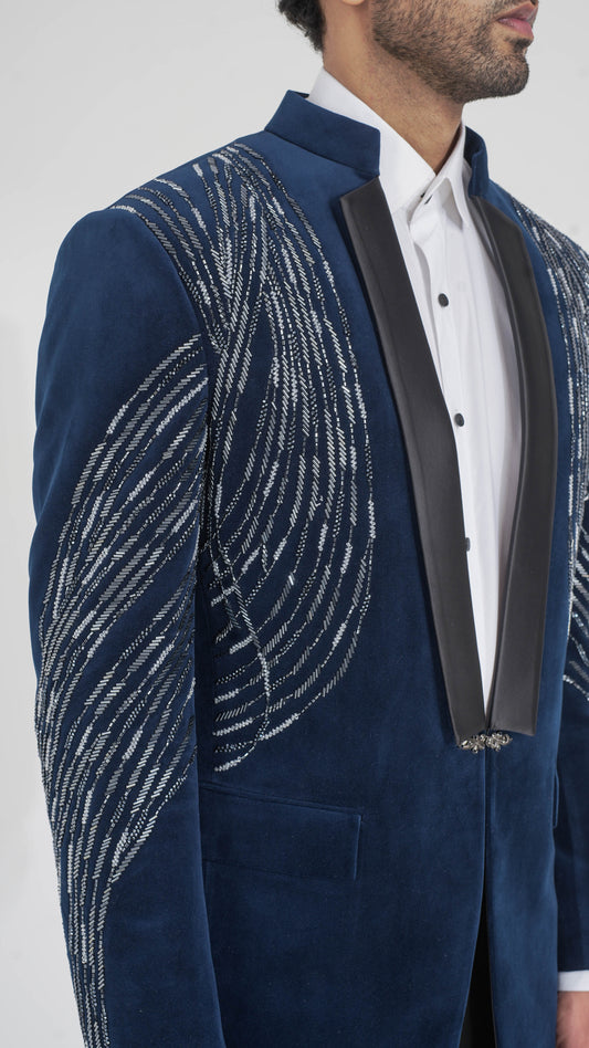 A close-up of a man wearing a navy blue velvet tuxedo jacket with silver embroidered wave patterns on the sleeves and front, paired with a white dress shirt and black satin lapels.