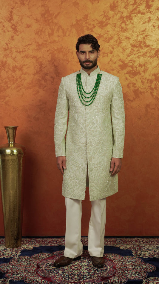 A man wearing a light green embroidered sherwani with white trousers and a green beaded necklace, standing against a textured orange wall next to a gold decorative vase.