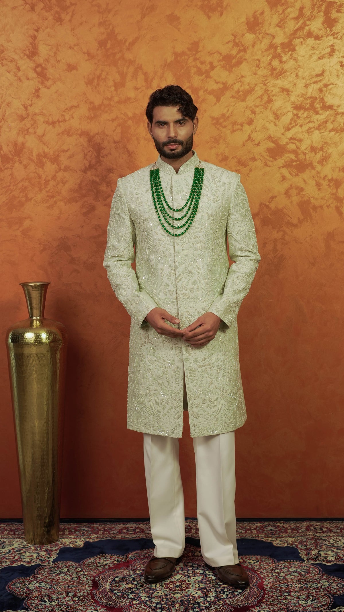 A man wearing a traditional Indian outfit, featuring a light green and white patterned sherwani with a double-layered green beaded necklace, standing in front of a textured orange-brown backdrop next to a tall gold vase.