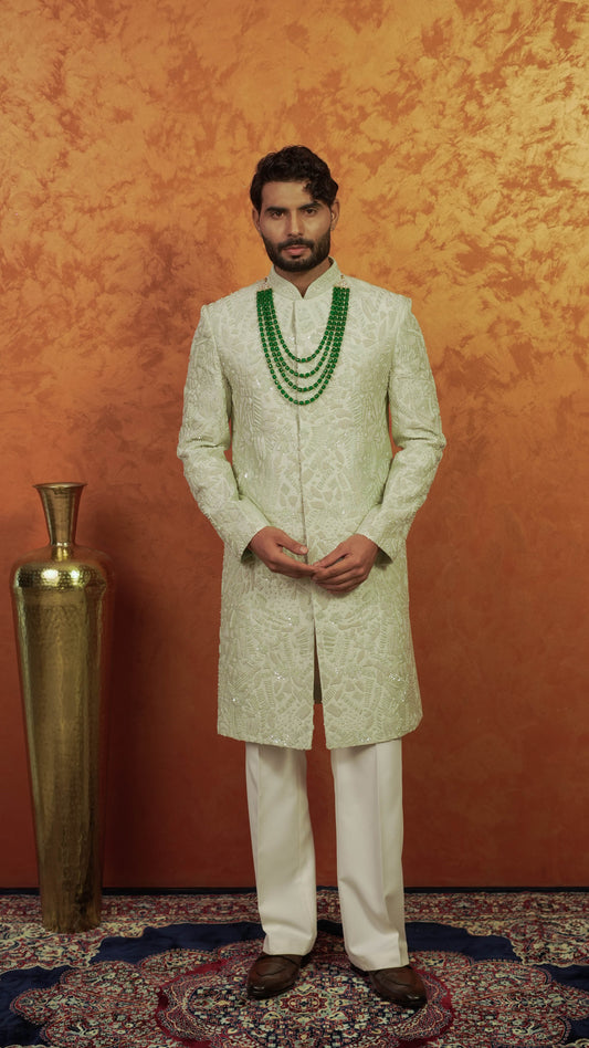 A man wearing a traditional Indian outfit, featuring a light green and white patterned sherwani with a double-layered green beaded necklace, standing in front of a textured orange-brown backdrop next to a tall gold vase.