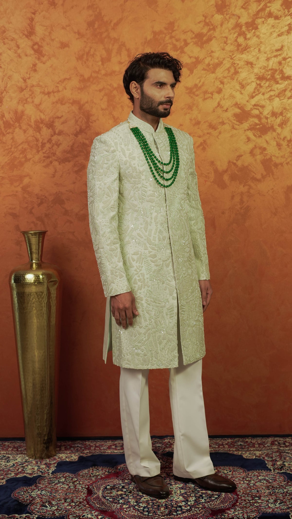 A man wearing a light green embroidered sherwani with white trousers and a green beaded necklace, standing in front of a textured orange wall next to a gold vase.