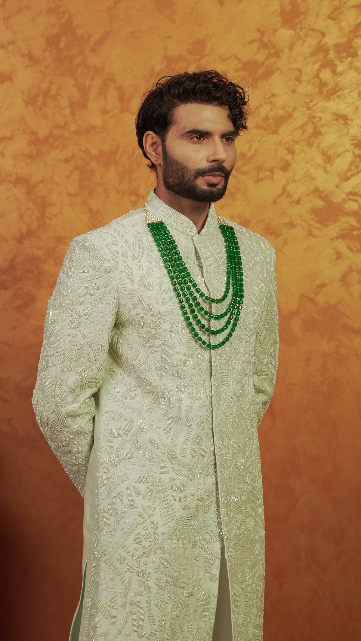 A man wearing a cream-colored embroidered sherwani with a multi-strand green beaded necklace, standing against a textured orange backdrop.