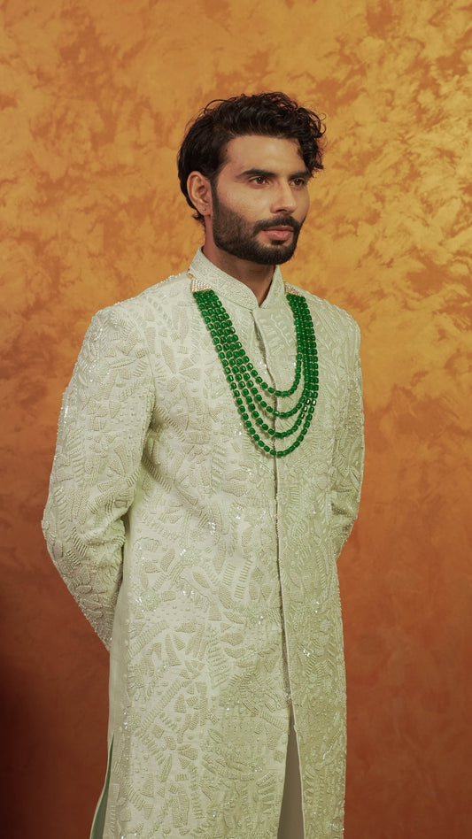 A man wearing a cream-colored embroidered sherwani with a multi-strand green beaded necklace, standing against a textured orange backdrop.