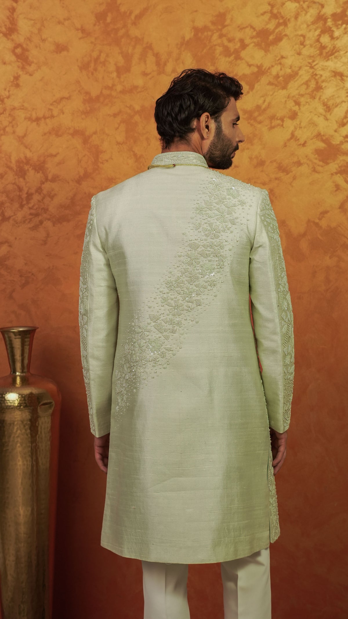 A man viewed from the back wearing a light sage green sherwani with intricate embroidery on the back, set against a textured orange background.