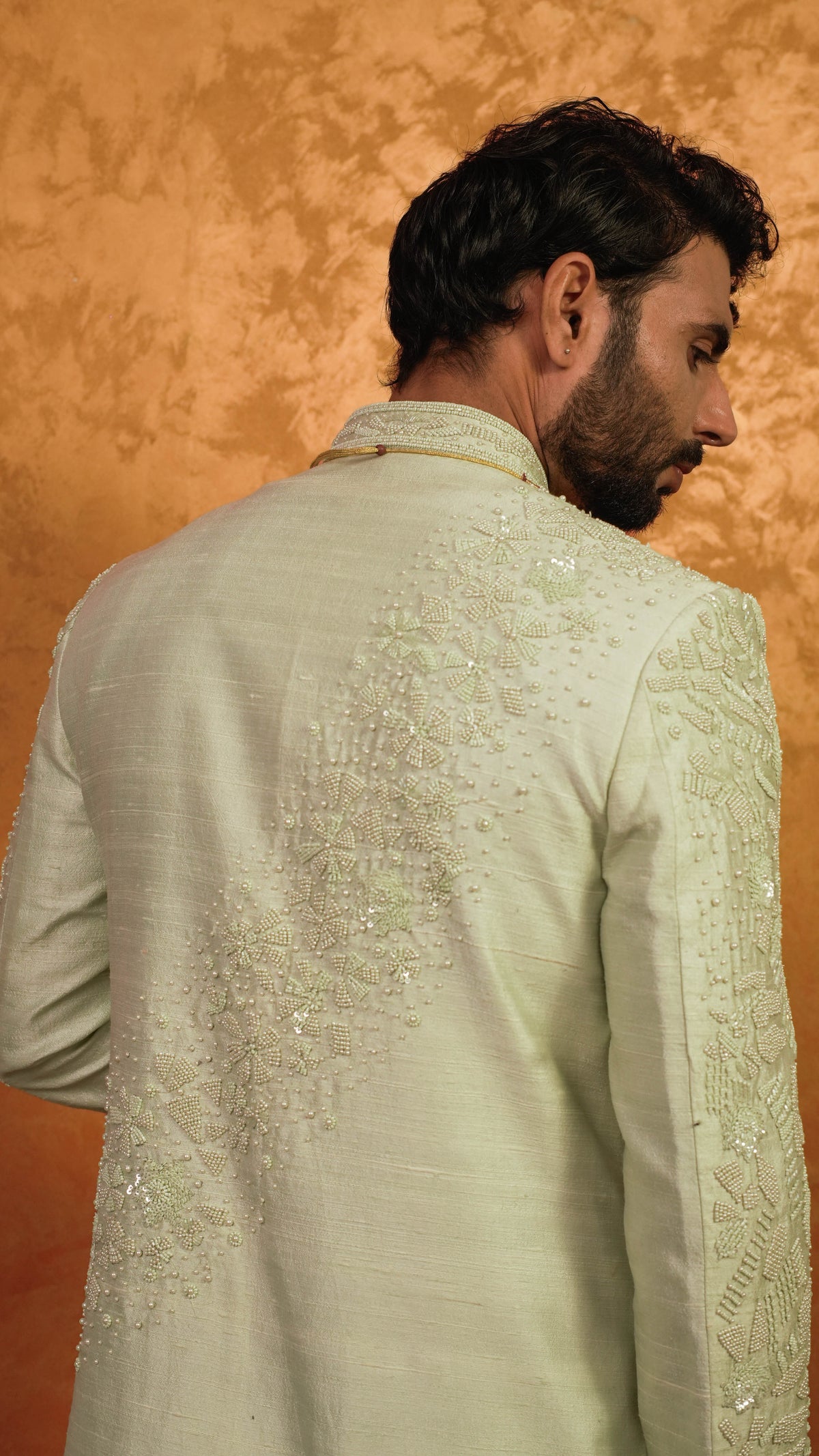 A man shown from the back wearing a light green sherwani jacket with intricate silver sequin embroidery on the back and sleeves, set against a textured warm orange-brown background.