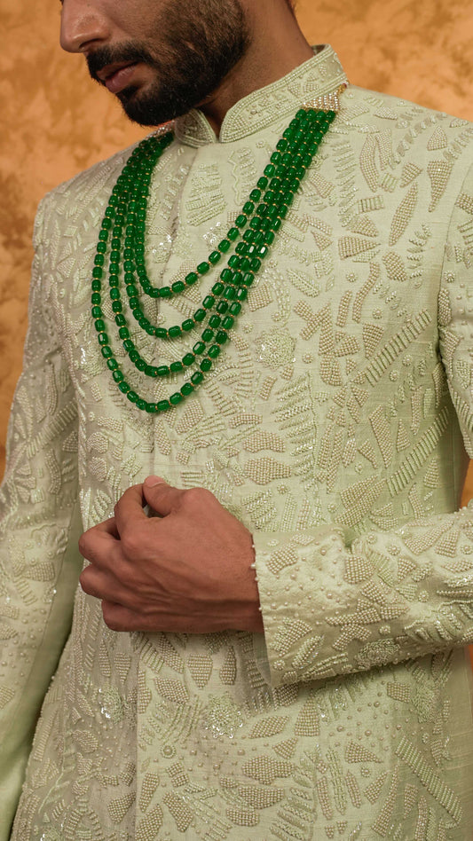A close-up of a man wearing a cream-colored embroidered sherwani with a matching mandarin collar, accessorized with a multi-strand green beaded necklace, against a textured gold background.
