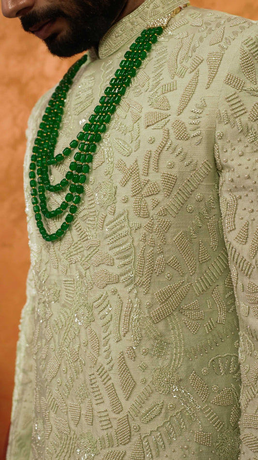 A close-up of a man wearing a light green embroidered sherwani with a double-row of vibrant green beaded necklaces.