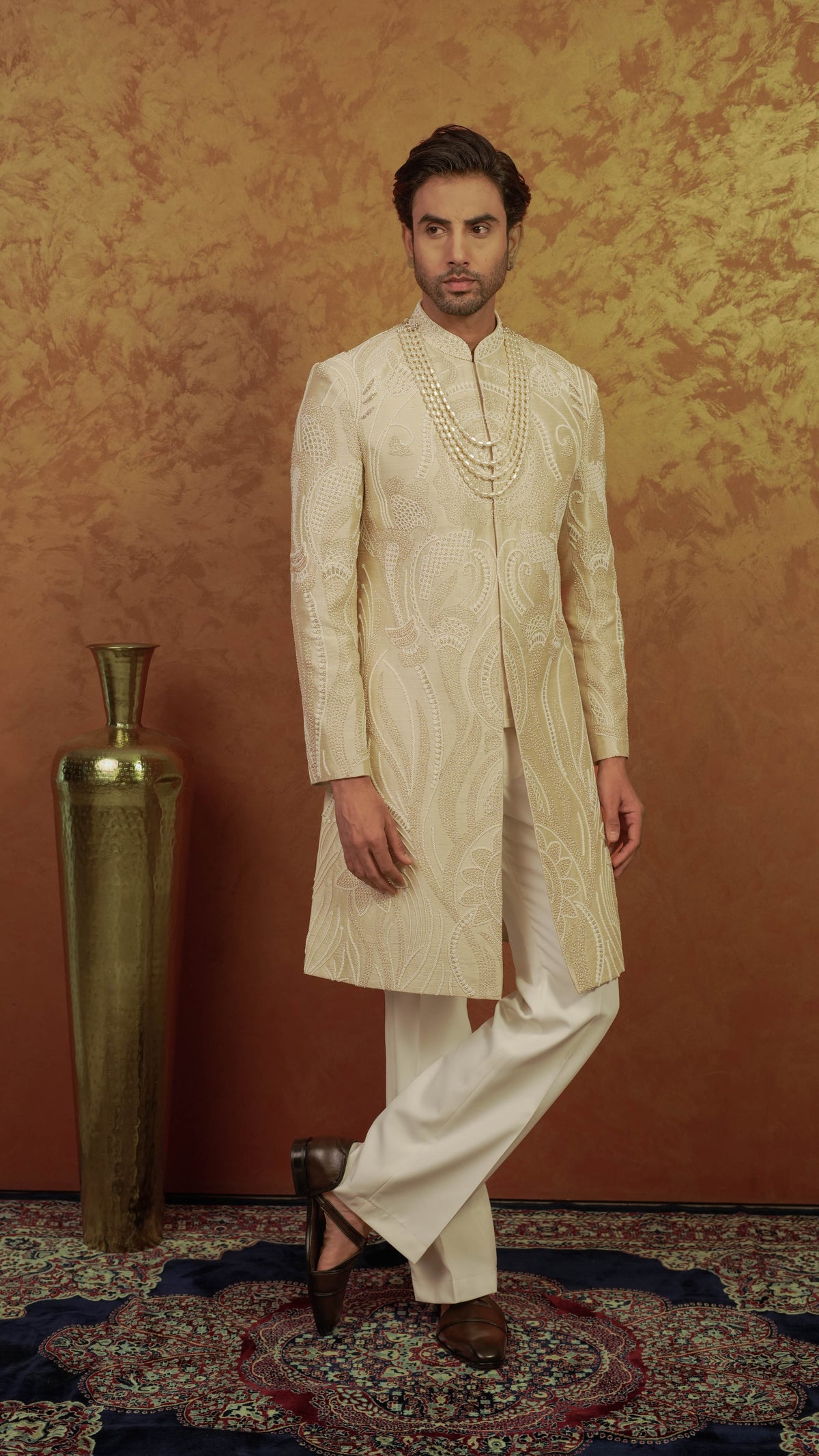 A man modeling a traditional Indian outfit consisting of a cream-colored embroidered sherwani jacket and matching trousers, standing in front of a textured golden-brown wall next to a tall brass vase.