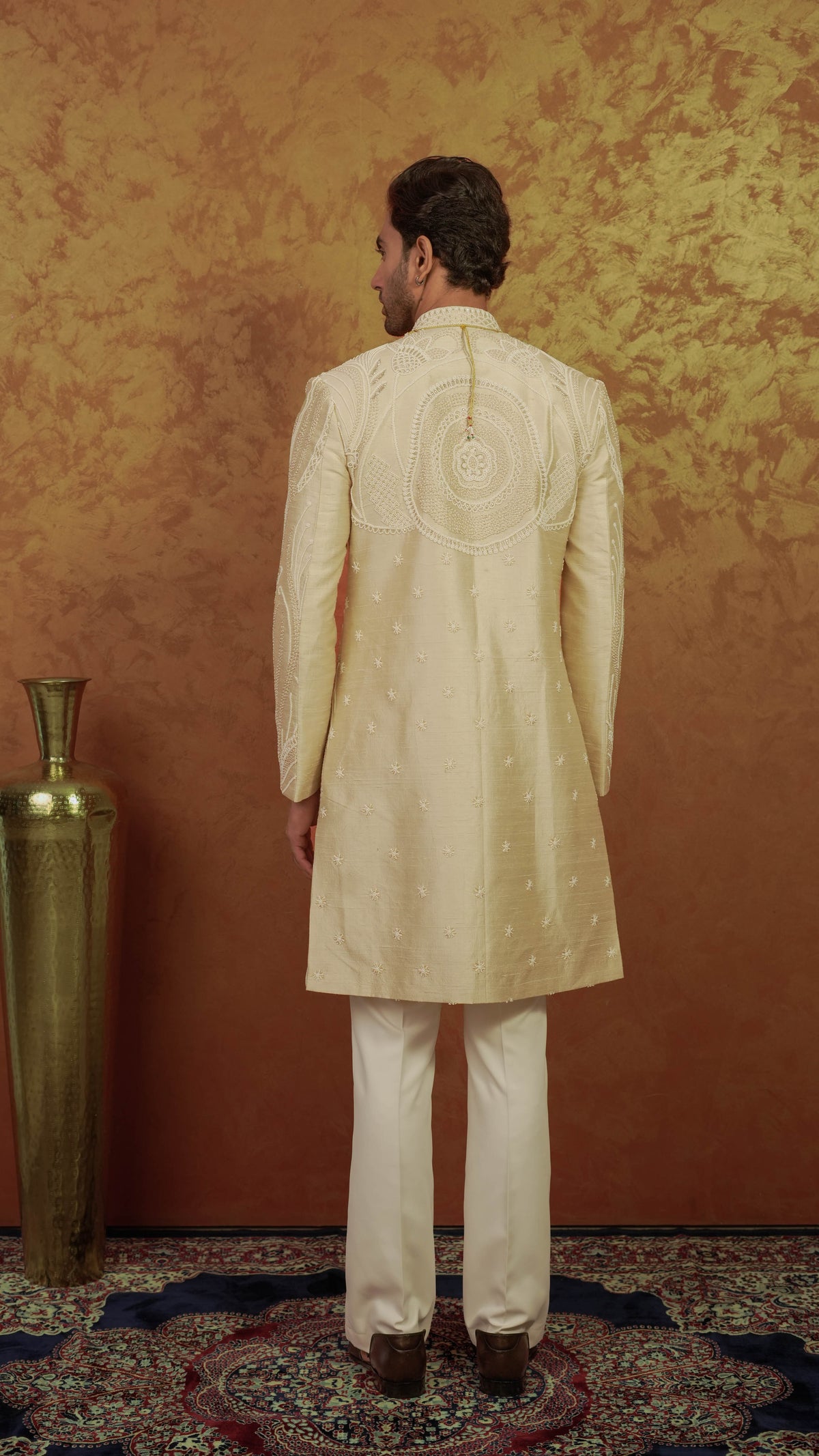 A man viewed from behind wearing a cream-colored sherwani with intricate embroidery on the back, paired with cream trousers, standing in front of a textured golden-brown wall.