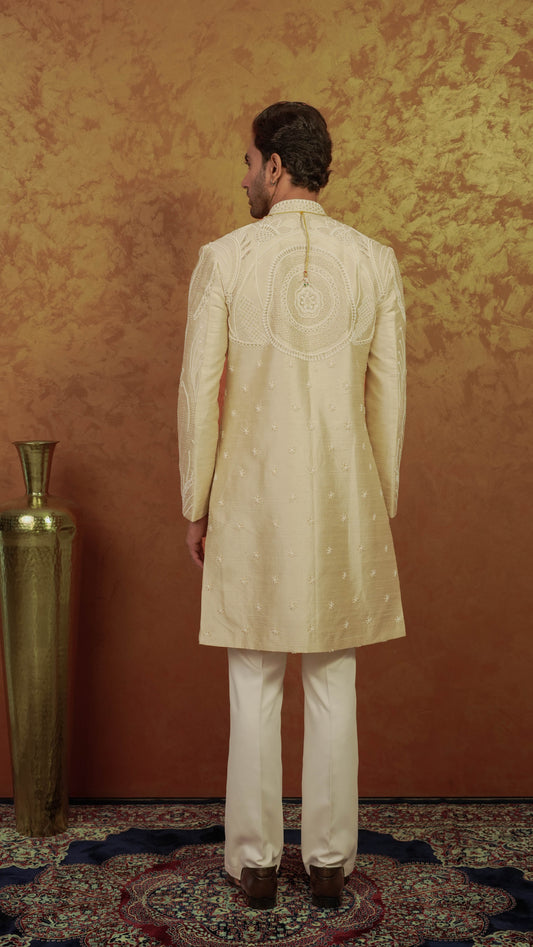 A man viewed from behind wearing a cream-colored sherwani with intricate embroidery on the back, paired with cream trousers, standing in front of a textured golden-brown wall.