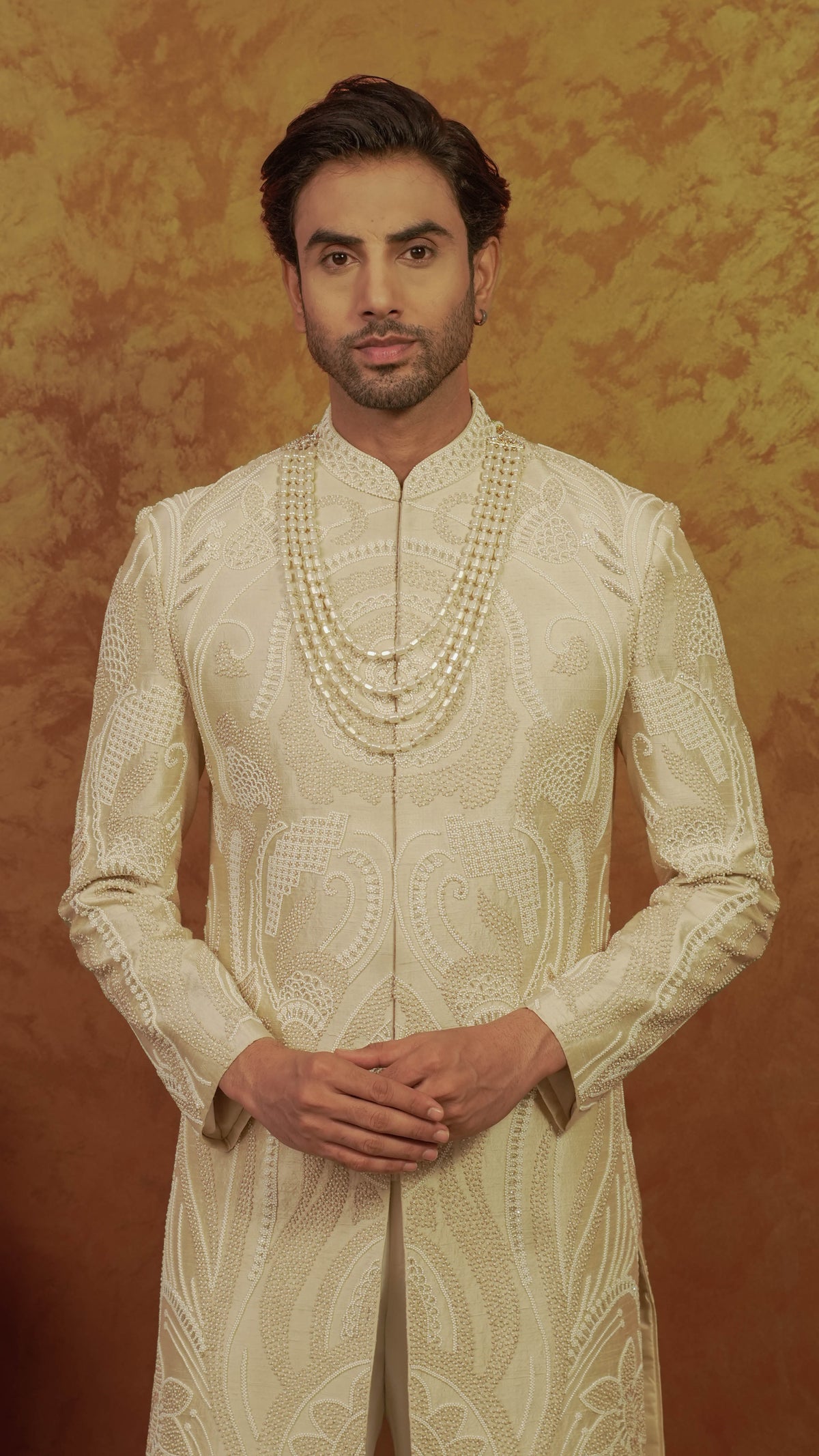 A man wearing a cream-colored, heavily embroidered sherwani with a matching multi-strand pearl necklace, standing against a textured golden-brown background.