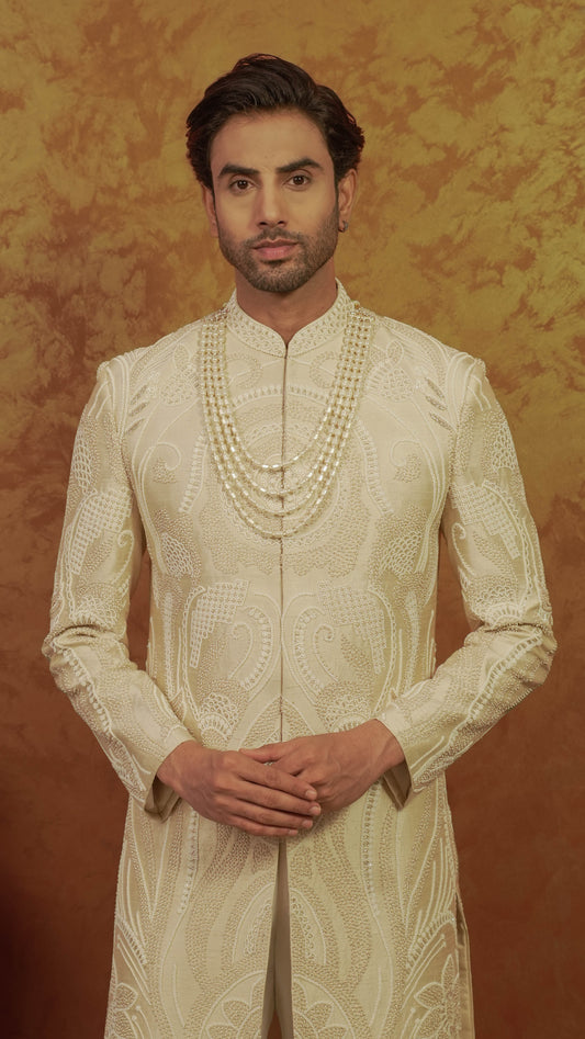 A man wearing a cream-colored, heavily embroidered sherwani with a matching multi-strand pearl necklace, standing against a textured golden-brown background.